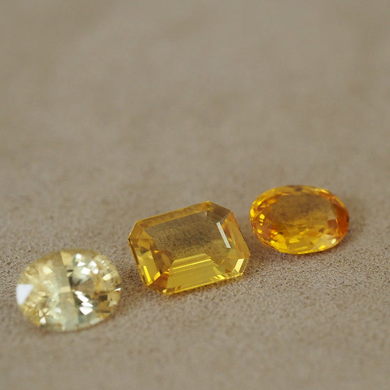 Three yellow gemstones of different cuts and facets arranged on a textured beige surface, including a round brilliant-cut stone, a rectangular emerald-cut stone, and an oval-cut stone.
