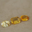 Three yellow gemstones of different cuts and facets arranged on a textured beige surface, including a round brilliant-cut stone, a rectangular emerald-cut stone, and an oval-cut stone.