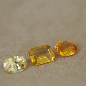 Three yellow gemstones of different cuts and facets arranged on a textured beige surface, including a round brilliant-cut stone, a rectangular emerald-cut stone, and an oval-cut stone.